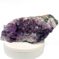 A natural amethyst crystal cluster with purple and lavender crystalline formations, displayed on a round white pedestal against a white background.