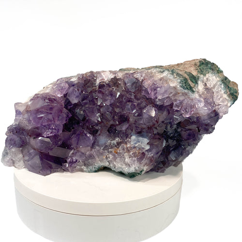 A natural amethyst crystal cluster with purple and lavender crystalline formations, displayed on a round white pedestal against a white background.