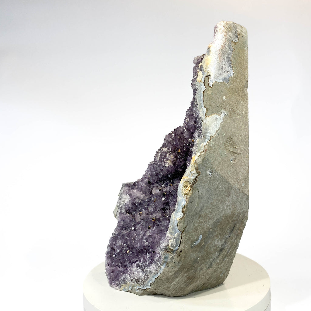 A natural amethyst geode specimen with a purple crystal cavity and gray outer matrix, displayed upright on a white pedestal against a clean white background.