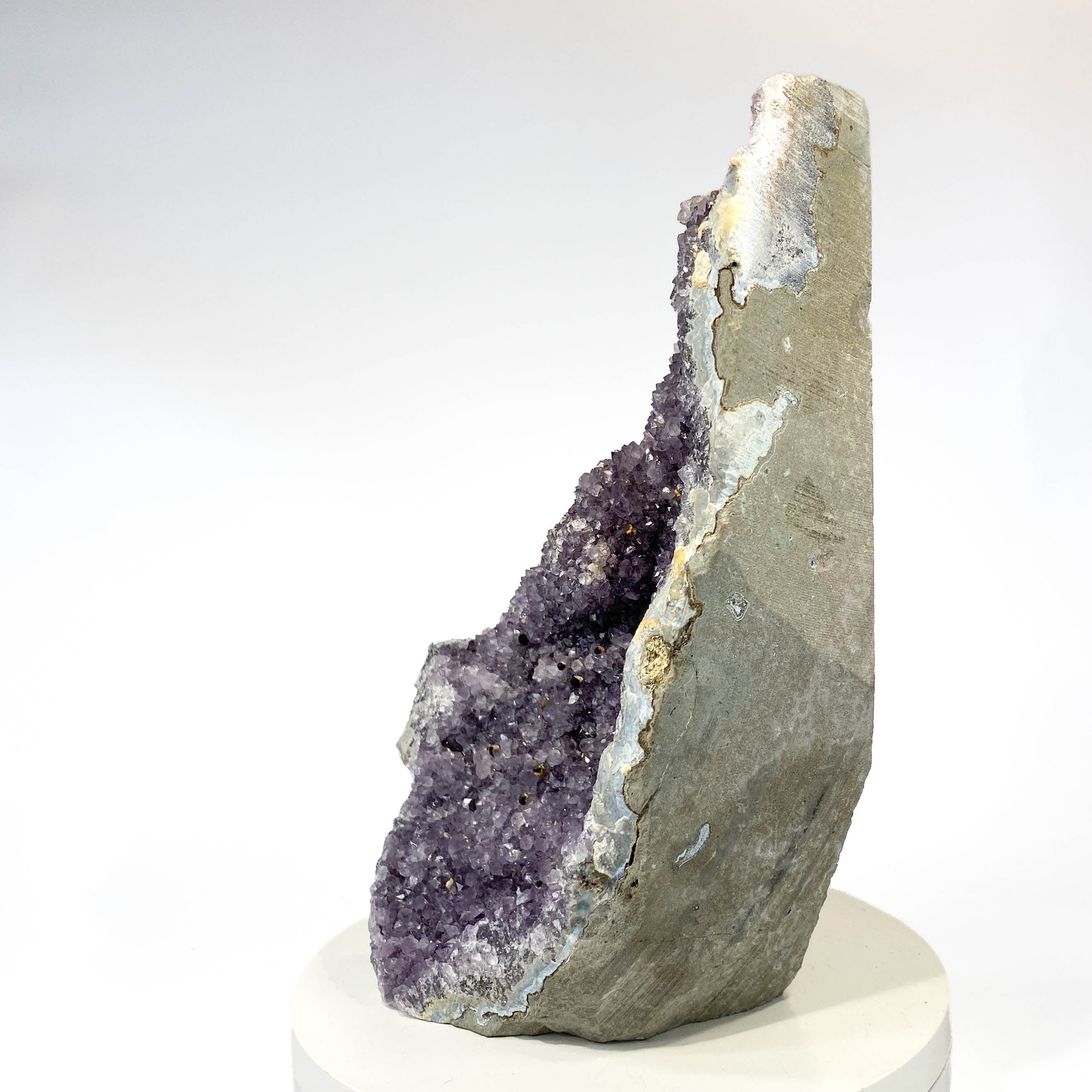 A natural amethyst geode specimen with a purple crystal cavity and gray outer matrix, displayed upright on a white pedestal against a clean white background.